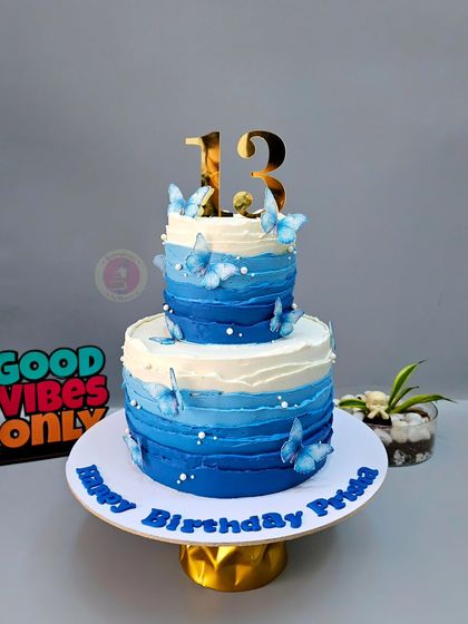 A stunning two-tier cake with a blue ombre watercolor effect, adorned with delicate blue butterflies for a 13th birthday.