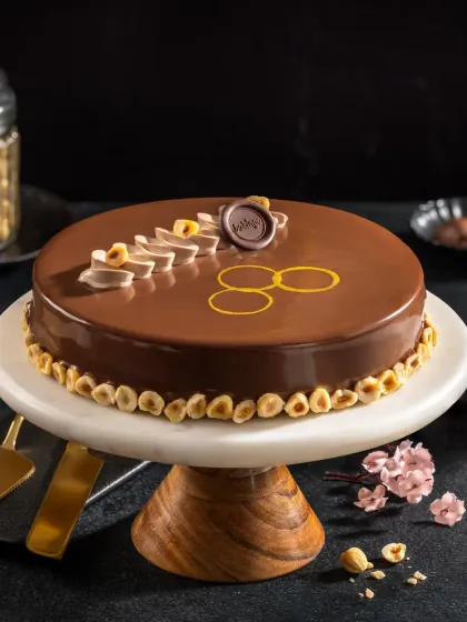 A glossy hazelnut chocolate cake with delicate piping and gold leaf accents, styled for a premium bakery's promotional materials.