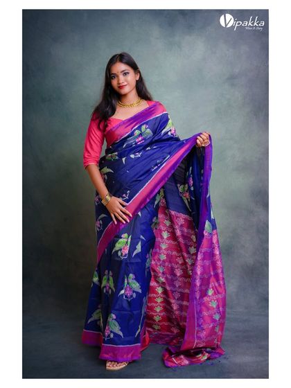 A classic studio shot for the brand Vipakka, featuring a model in a rich blue and pink printed saree. The mottled grey background adds a touch of texture without distracting from the product.