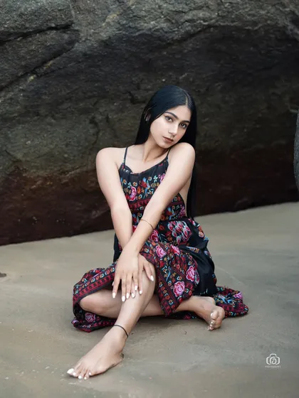 A simple and beautiful portrait of a model in a floral dress, sitting on the sand. The focus is on her relaxed pose and the natural beauty of the beach setting.