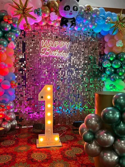 A vibrant first birthday party setup featuring a full balloon arch with a panda theme, a sparkling silver sequin wall, a "Happy Birthday" neon sign, and a large light-up marquee number '1'.