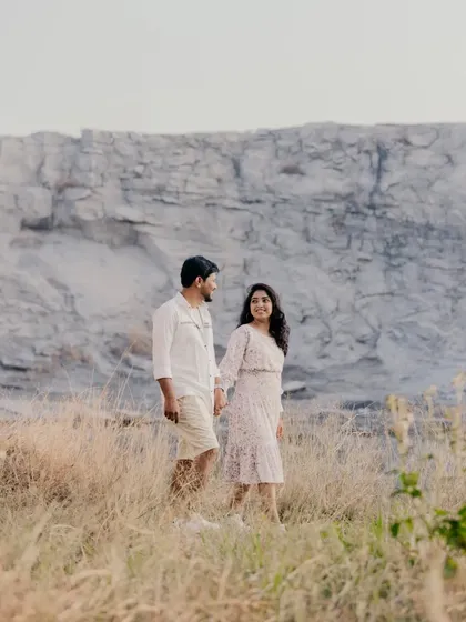 A candid shot of a couple walking through a field of tall, dry grass, with a massive rock formation in the background.