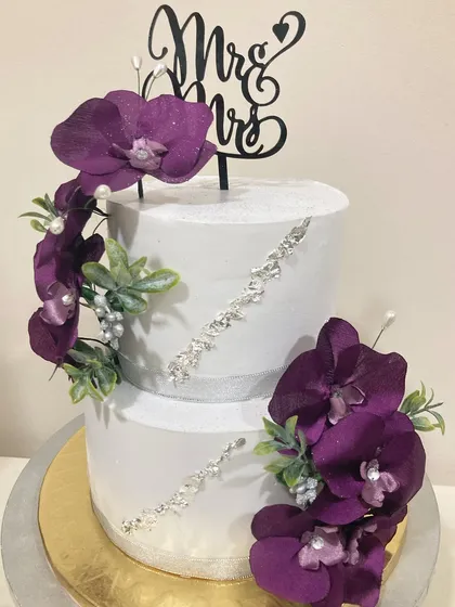 A modern and luxurious two-tier wedding cake. The crisp white fondant is accented with diagonal crystal detailing and cascades of deep purple orchids, complete with a "Mr & Mrs" topper for a sophisticated finish.