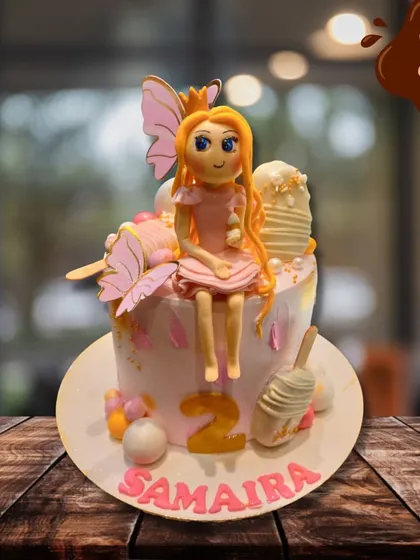 A sweet fairy-themed cake for a second birthday. This design features a hand-sculpted fairy with butterfly wings sitting atop a pink cake, decorated with mini cakesicles and the name Samaira.