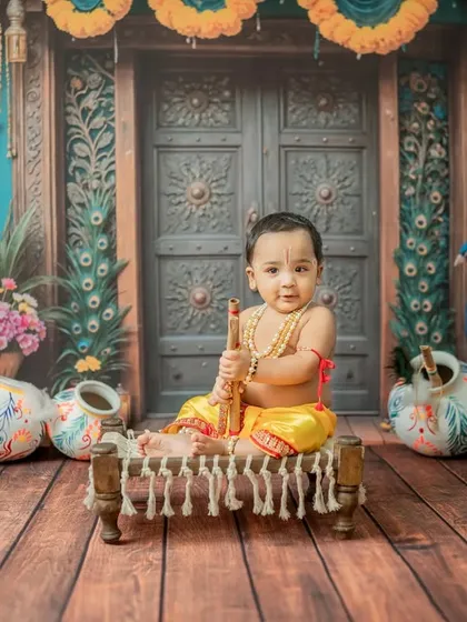 A simple, direct portrait of baby Krishna on the cot, holding his flute, with the ornate door in the background.