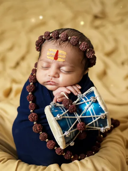 A newborn wrapped in blue, holding a tiny damru. This Shiva-themed shoot was done at the client's home, showing our ability to create unique setups anywhere.