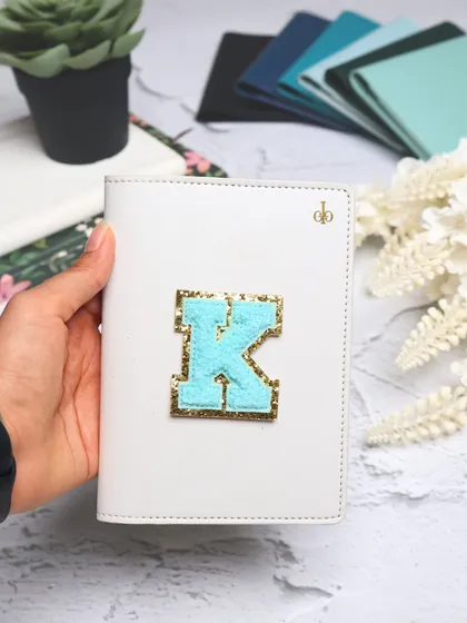 A close-up of our white passport cover, personalized with a light blue and gold glitter chenille "K" patch. You can choose from over 12 cover colors and any initial.