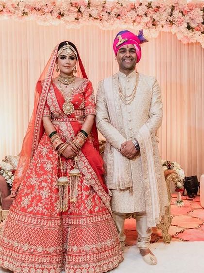 A beautiful couple poses against a soft and romantic wedding backdrop. The stage features a wall of pink and white flowers, creating a delicate and pretty setting for an indoor ceremony.