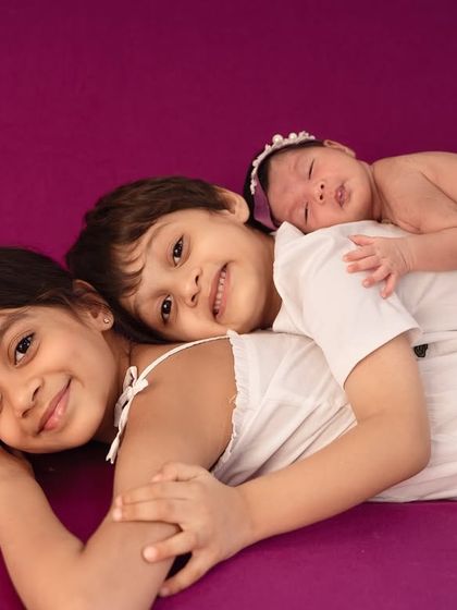Three siblings snuggled together, with the newest addition resting peacefully. The vibrant purple backdrop adds a pop of color to this sweet and loving newborn and sibling portrait.