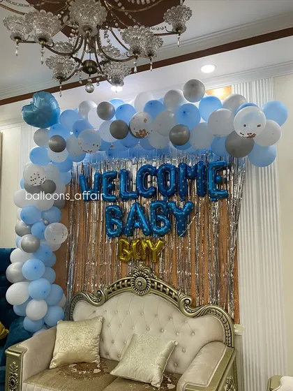 A 'Welcome Baby Boy' setup in a living room, featuring a balloon arch against a silver shimmer curtain and an elegant sofa.