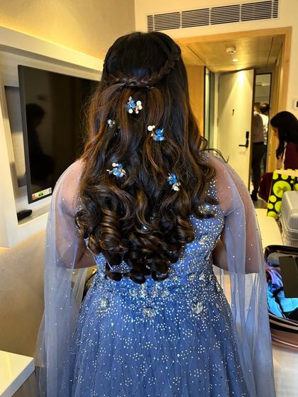 A princess-inspired hairstyle for an engagement. This half-updo features a neat braid across the crown and voluminous curls adorned with delicate blue floral accessories to match her gown.