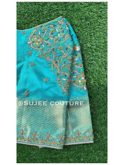 A close-up of a teal blouse sleeve. The design features a beautiful tree-of-life pattern embroidered with green and gold zardozi work on a contrasting silk brocade fabric.