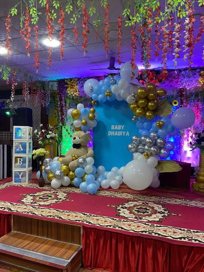 A stage setup for a baby boy's celebration. The decor includes a blue backdrop, a balloon arch in blue, white, silver, and gold, and a large teddy bear as the centerpiece.