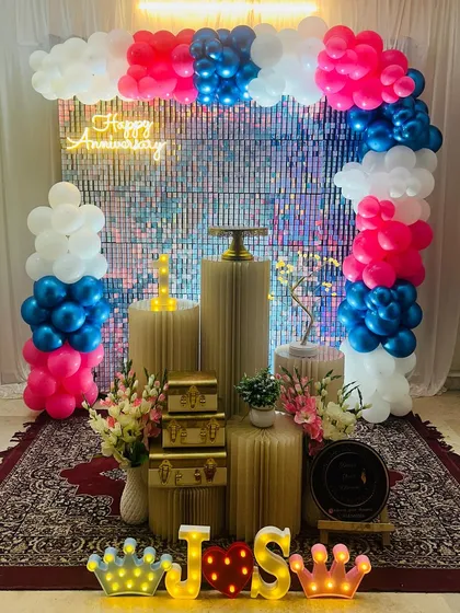 A glamorous anniversary party decoration with a sparkling silver shimmer wall. The setup is completed with a colorful balloon arch, a 'Happy Anniversary' neon sign, and decorative plinths for props.