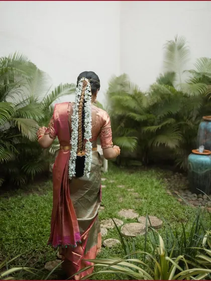 A back shot of a South Indian bride in a beautiful silk saree, her long braid adorned with traditional flowers, walking through a garden.