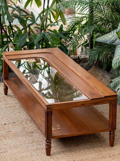 The Kulam center table, inspired by the serene temple ponds of Kerala. The design features a glass top over a wooden base, with brass details beneath the surface that depict elements like lotus leaves and fish, creating a miniature aquatic scene.