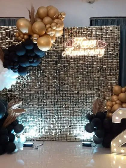 Celebrating a 25th anniversary in style at home. This setup features a gold sequin wall, a "Happy Anniversary" neon sign, and a black, white, and gold balloon arrangement.