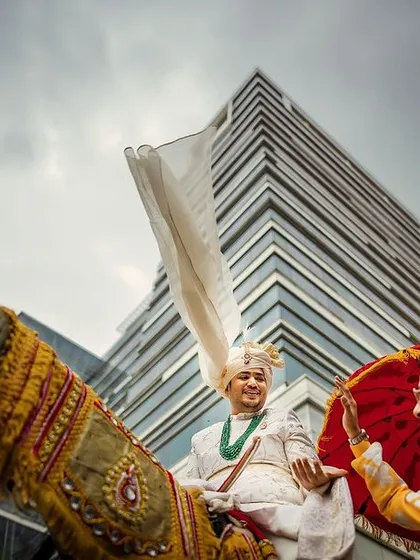 A dynamic low-angle shot of the groom's baraat entry on a horse, set against a modern cityscape. This image blends tradition with a contemporary, energetic filming style.