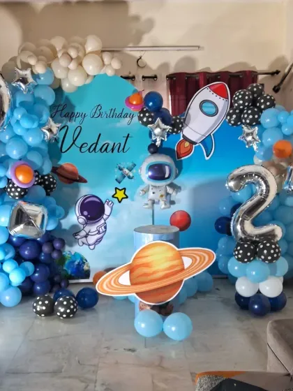 An exciting space-themed birthday decoration for a second birthday. This setup includes a custom backdrop, astronaut and rocket cutouts, planet props, and a balloon garland in different shades of blue.