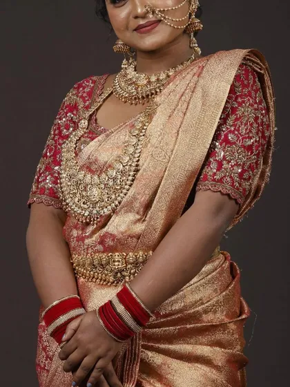This is a classic South Indian muhurtham look. The rich red and gold Kanchipuram silk saree is paired with a custom-designed blouse featuring intricate zardozi and maggam embroidery, creating a timeless bridal outfit available for rent.