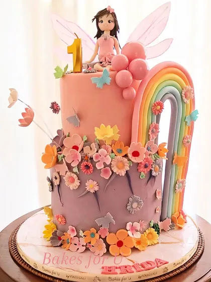 A magical two-tier fairy and rainbow cake for a first birthday. This enchanting design features a fairy figure, colorful flowers, and a beautiful fondant rainbow arch.