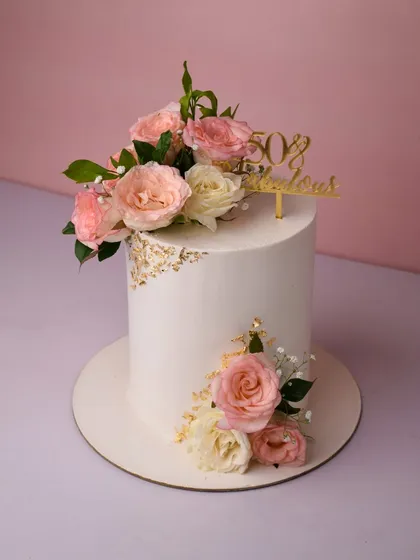 An elegant tall cake for a "50 & Fabulous" celebration. The smooth white finish is accented with delicate gold leaf and a beautiful arrangement of fresh pink and white roses, creating a sophisticated and timeless design.