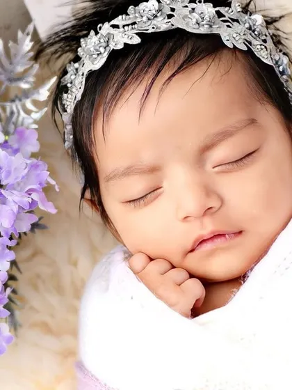 A close-up of a baby girl in a beautiful silver-beaded headband, resting against purple flowers.