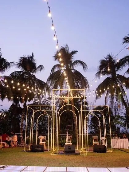 A modern Sangeet setup with a checkered dance floor and a minimalist gold arch structure. The view captures the open-air feel with palm trees and string lights against the twilight sky.