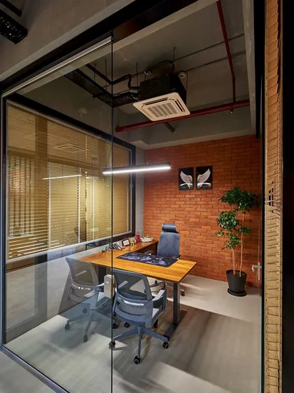A private executive cabin featuring an exposed brick wall, a sleek wooden desk, and a potted plant. The glass partition ensures the space feels connected to the larger office.