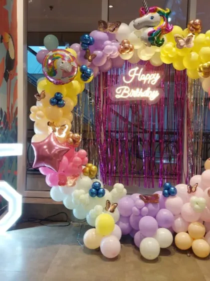 A magical unicorn-themed party for a 5th birthday. This vibrant setup includes a circular balloon arch in pastel rainbow colors, a sparkling pink fringe backdrop, a 'Happy Birthday' neon sign, and a large light-up number '5' for a truly enchanting celebration.