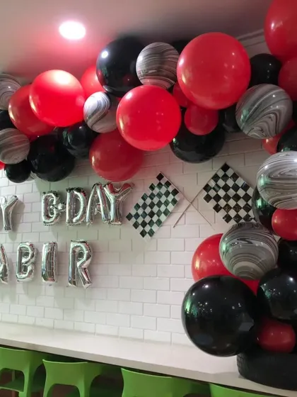 A race car-themed birthday party for Kabir. The setup includes a balloon arch with red, black, and marbled agate balloons, checkered flags, and silver foil letters against a white brick wall.