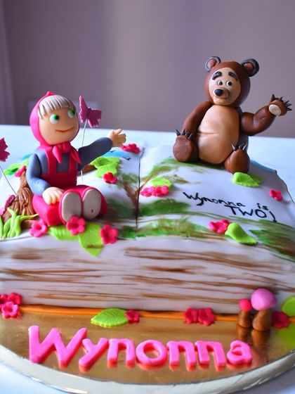 A storybook cake come to life for a 2nd birthday. This cake is shaped like an open book and features adorable, hand-sculpted fondant figures of Masha and the Bear in a forest setting.