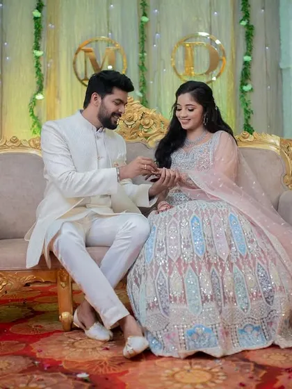 The groom places the ring on the bride's finger as they sit on an ornate sofa during their engagement.