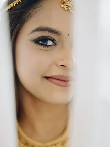 An artistic and intimate portrait of a bride. This close-up shot peeking through the veil highlights the precision of the winged eyeliner and the soft, matte finish of the lips, creating a captivating and mysterious look.