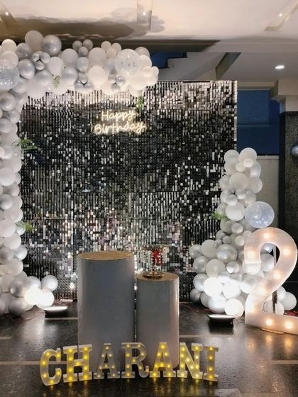 A stylish 21st birthday setup with a black shimmer wall, an all-white balloon arch, and illuminated marquee numbers and letters spelling out a name.