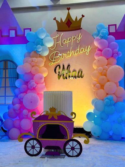 A charming princess carriage theme for Vihaa's 5th birthday. The backdrop includes a neon "Happy Birthday" sign, a crown, and a cute carriage prop, all surrounded by pastel pink, orange, and blue balloons.