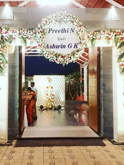 This grand entrance to a wedding hall is framed by a lush floral arch of white and red roses. A personalized welcome sign with the couple's name is placed within a floral wreath at the top of the arch.