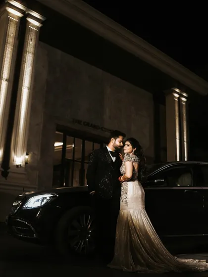 A glamorous outdoor portrait of the couple on their reception night. Posed in front of a luxury car and a grand, well-lit building, this shot has a sophisticated and high-fashion feel.