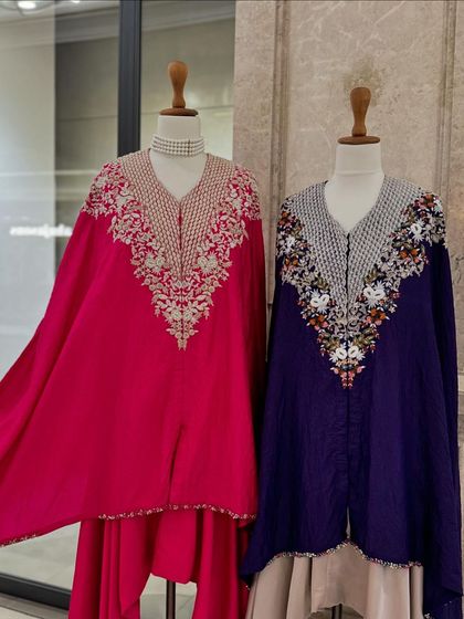 A display of two kaftan-style outfits with cape-like sleeves, in vibrant fuchsia and royal blue, featuring heavily embroidered necklines.