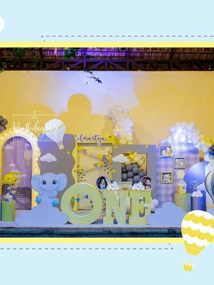 A cheerful and whimsical first birthday celebration with a hot air balloon and baby elephant theme. The complete stage setup features large 'ONE' block letters, custom backdrops, and a soft color palette of yellow, grey, and blue.