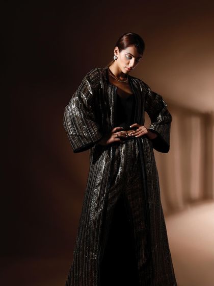 A powerful, full-length shot of a model in a shimmering, floor-length jacket. The dramatic, low-key lighting creates a sense of mystery and luxury.