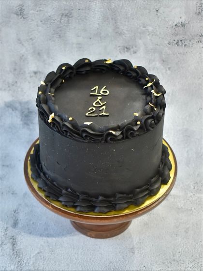 The top view of the black ganache cake, showing the "16 & 21" inscription for a joint sibling birthday, with delicate gold leaf accents on the piped border.