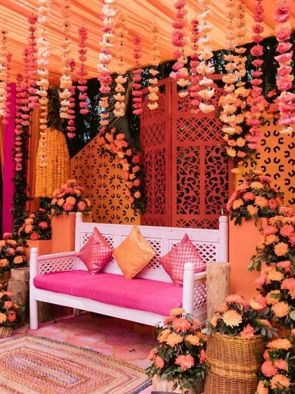A cozy and vibrant photo corner at a mehendi. A white bench with pink cushions is surrounded by orange floral arrangements and a patterned backdrop, perfect for guest photos.