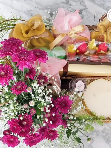 A dry fruit and sweets hamper bursting with color, featuring bright pink chrysanthemums, a large scented candle, and other festive treats.