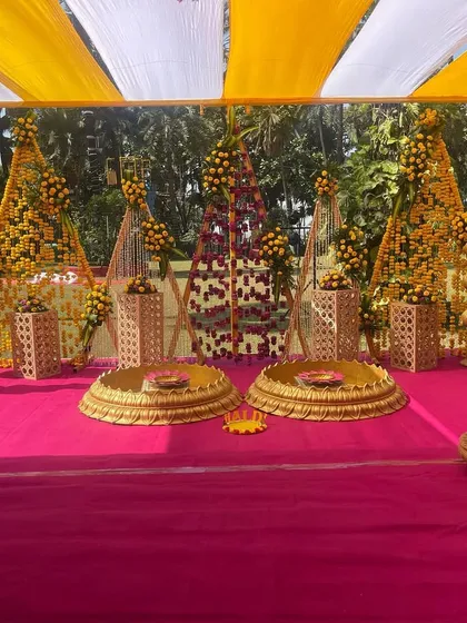 A traditional Haldi mandap setup on a pink stage. The decor features triangular backdrops made of marigolds and brass temple pillars, blending modern shapes with classic elements.