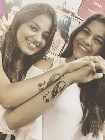 Sisters with matching "Dad Mom" infinity feather tattoos on their forearms.