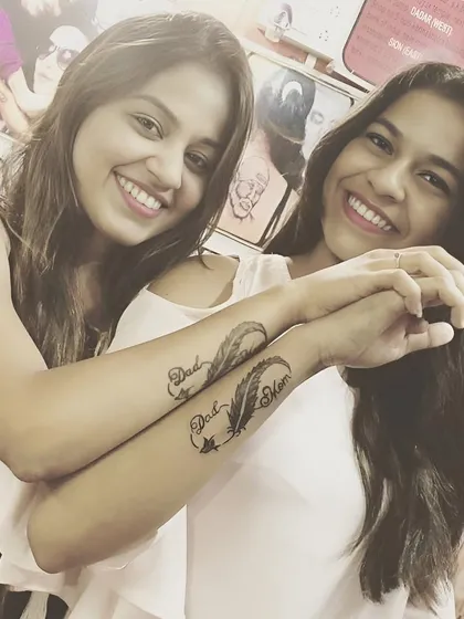 Sisters with matching "Dad Mom" infinity feather tattoos on their forearms.