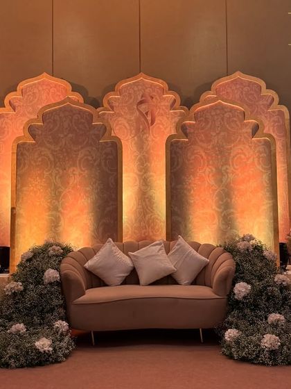 An elegant stage for a Nikah or reception, with a backdrop of Mughal-style arches and a sofa surrounded by a cascade of baby's breath and pink flowers.