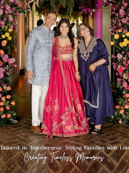 Tailored in togetherness. Here, the bride stands with her parents, showcasing a beautiful blend of colors and styles for the Sangeet. The bride's hot pink lehenga is complemented by her father's soft blue kurta and her mother's deep navy outfit, creating a timeless family portrait.
