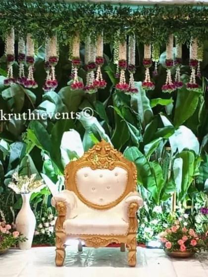 This is a classic South Indian decor theme featuring a backdrop made entirely of fresh banana leaves. The hanging jasmine garlands and ornate gold and white chair create a look that is both traditional and incredibly elegant.
